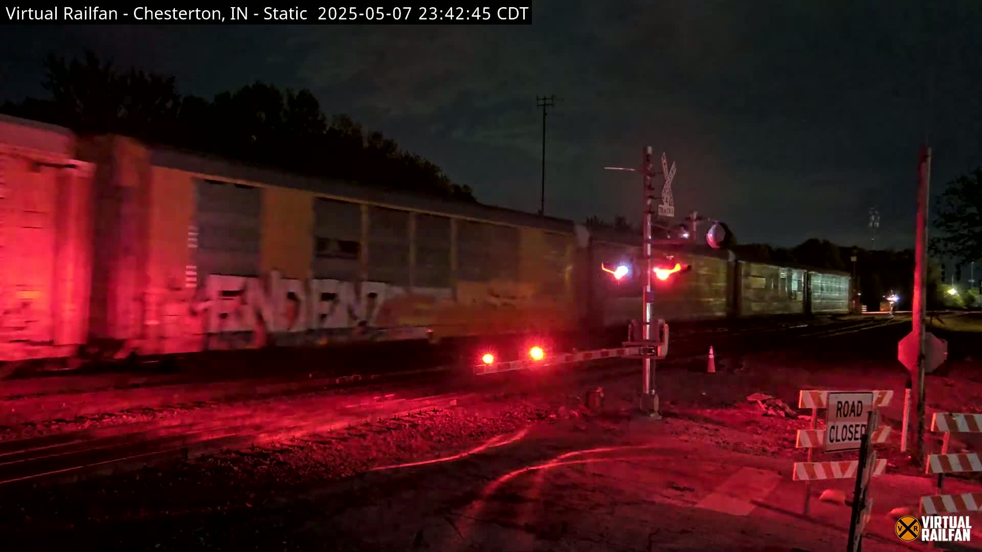 A long freight train with graffiti passes a nighttime railroad crossing with the gates down and red flashing lights illuminating the scene.