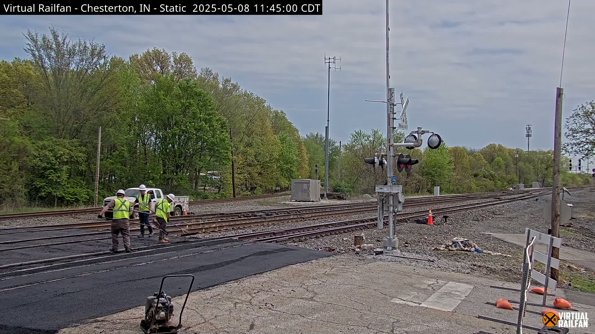 Several workers in bright safety vests are smoothing fresh asphalt at a railroad crossing under a partly cloudy sky.