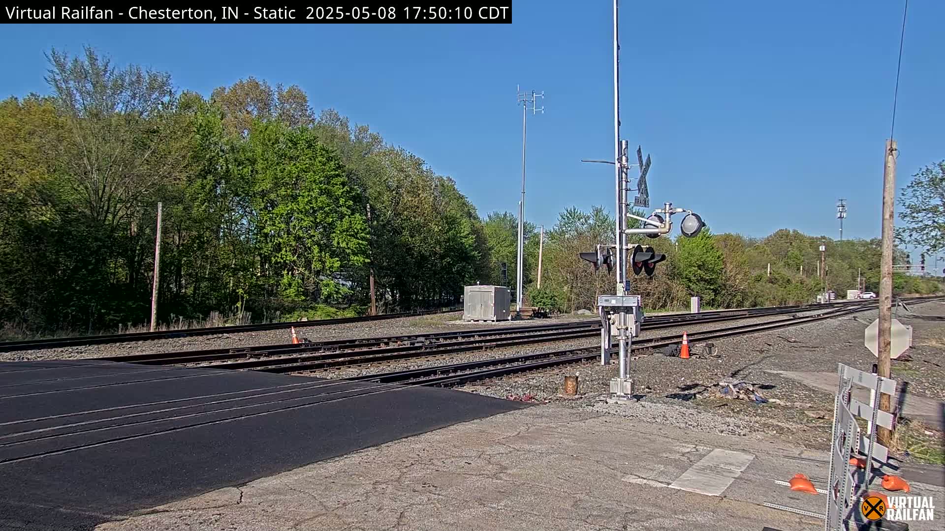 A railroad crossing with freshly paved asphalt shows two sets of train tracks extending into a background of green trees under a clear blue sky.