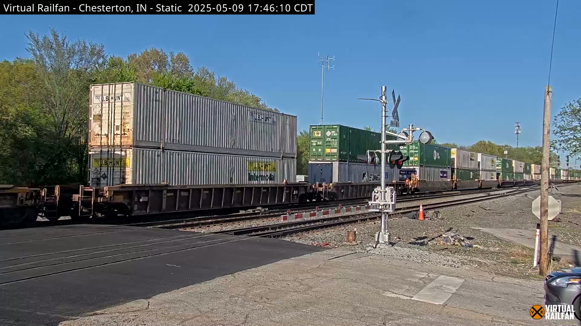 A long freight train carrying stacked shipping containers passes a railroad crossing on a sunny day.