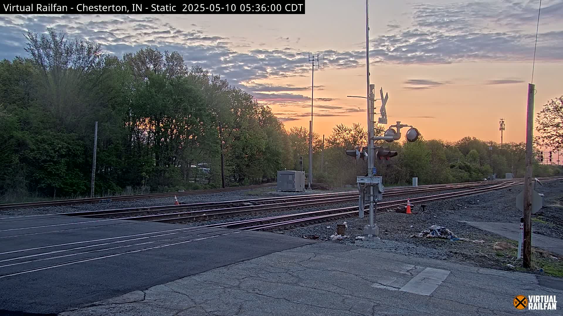 A railroad crossing at sunset, with multiple sets of train tracks intersecting a paved road, surrounded by trees under a partly cloudy sky.
