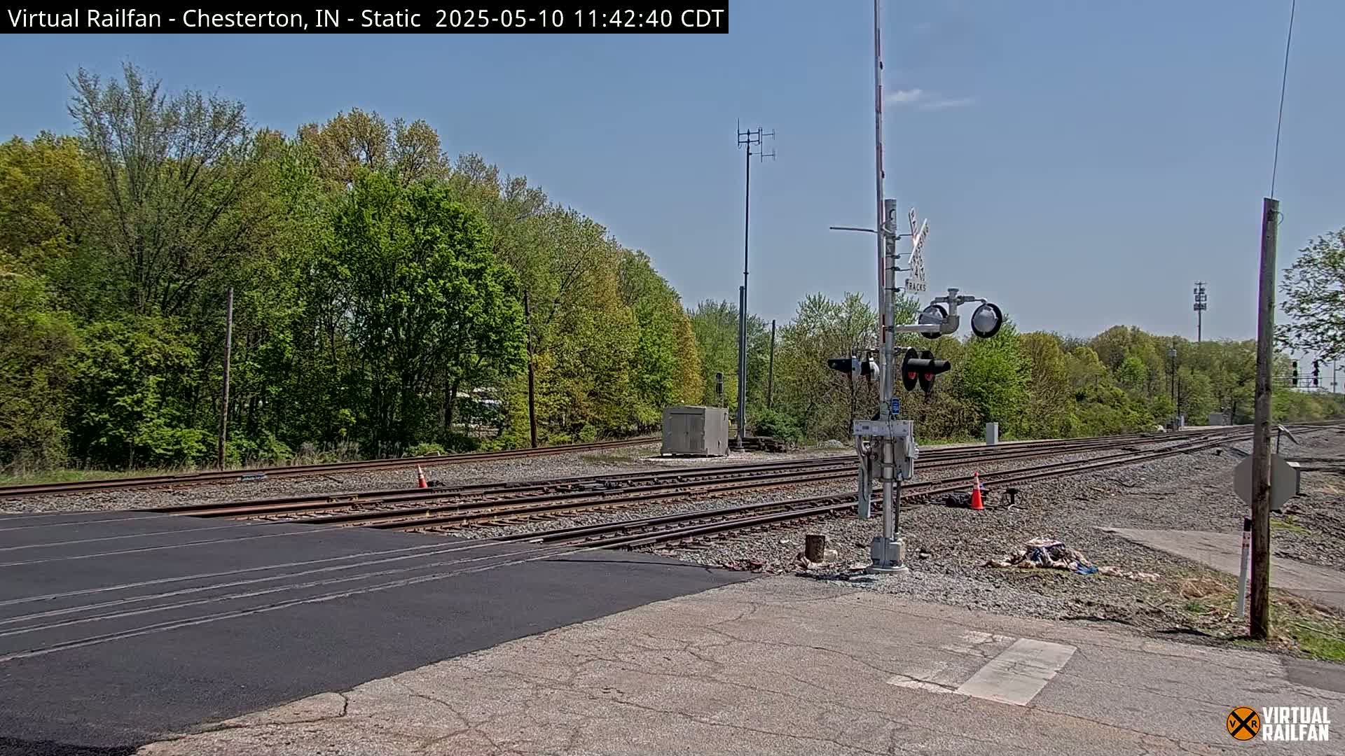 A railroad crossing with multiple tracks is shown on a sunny day, surrounded by lush green trees.