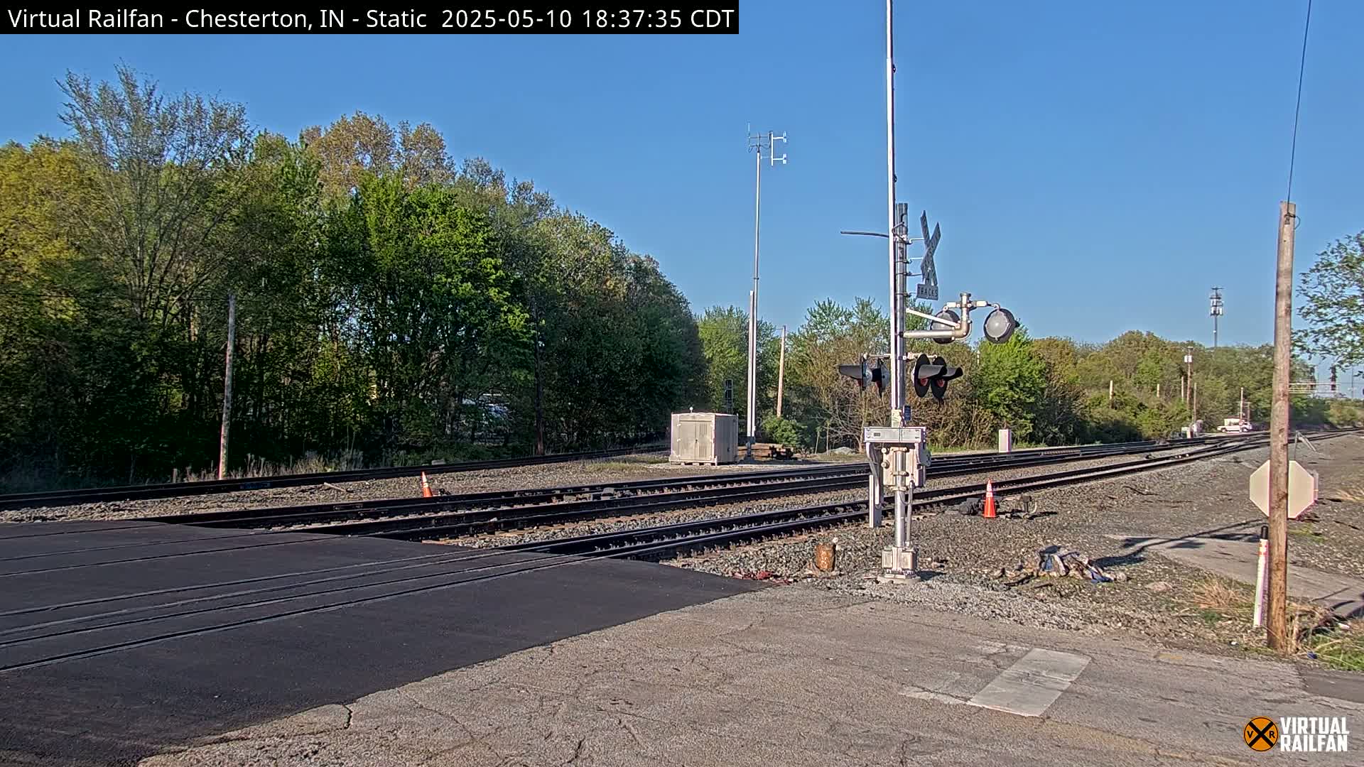 A railroad crossing with new asphalt, train tracks, and signal equipment is visible under a clear blue sky, surrounded by green trees.