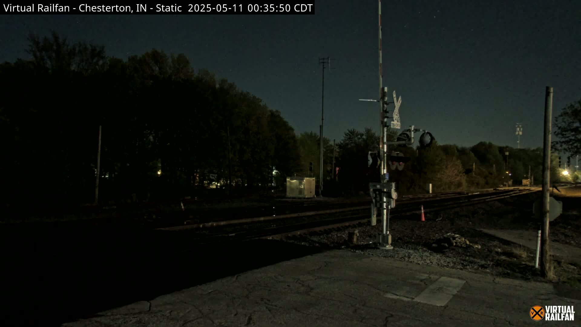 A nighttime view of a railroad crossing with train tracks extending into a dark, tree-lined area under a clear, starry sky.