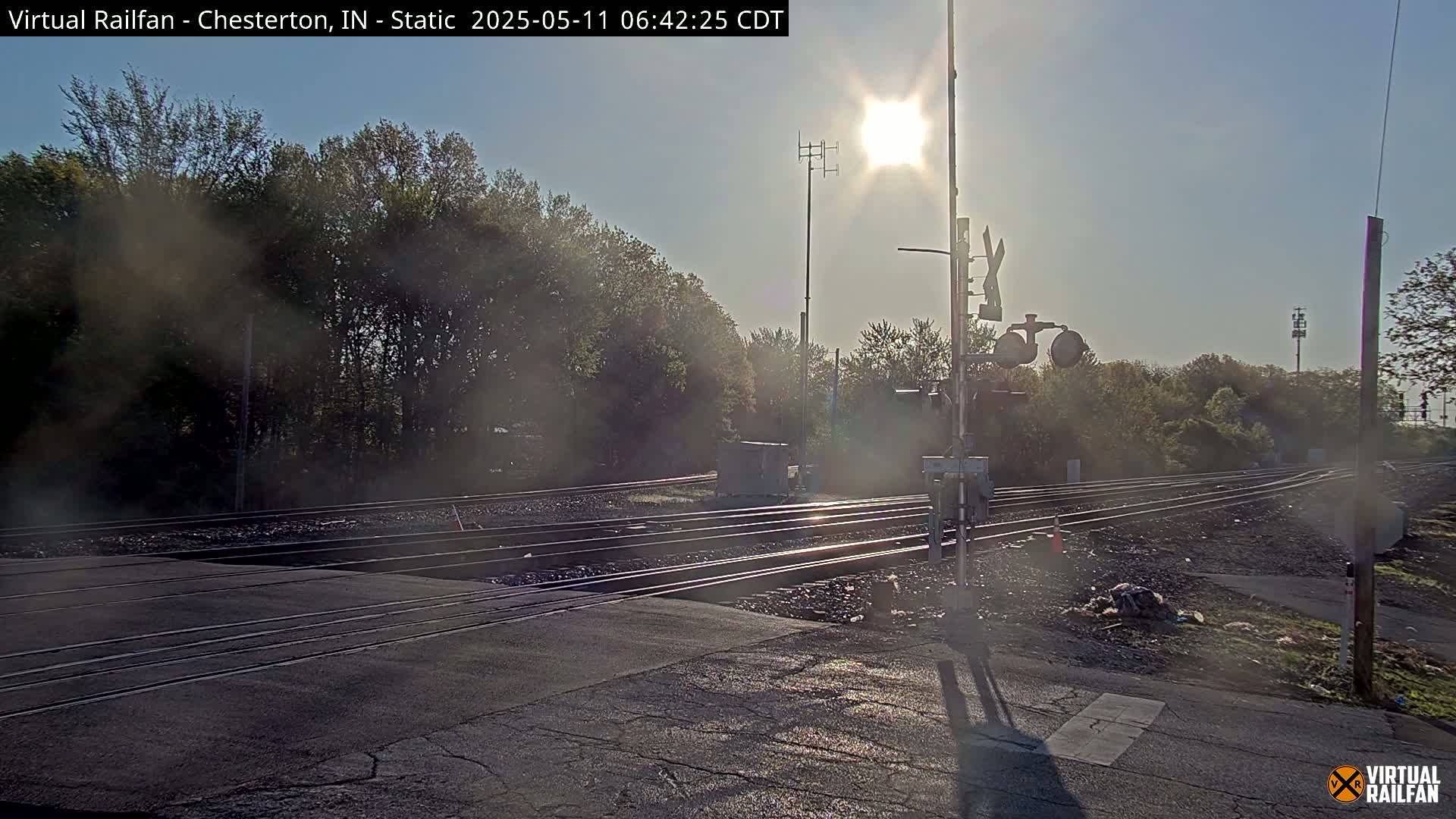 A sunny day at a railroad crossing shows multiple train tracks intersecting a road, with trees lining the background.