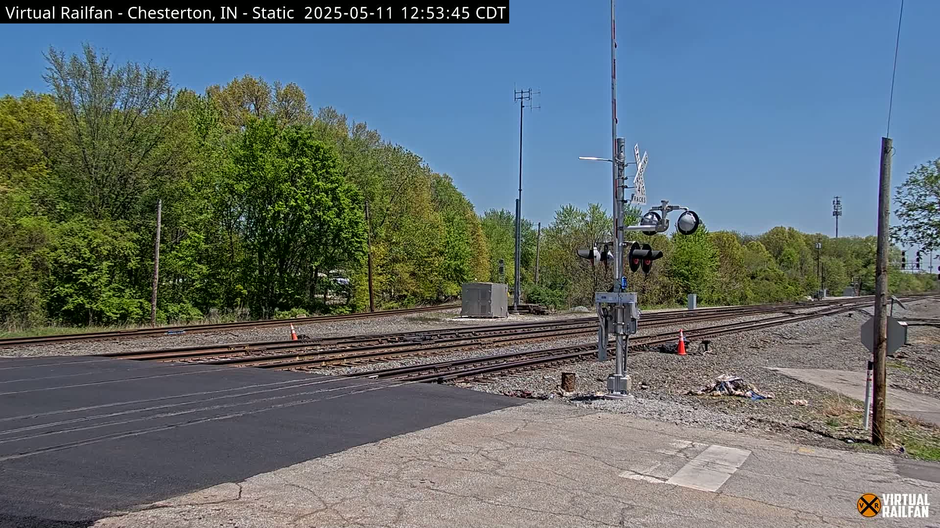 A railroad crossing with multiple tracks, crossing signals, and a surrounding area of green trees under a clear blue sky.