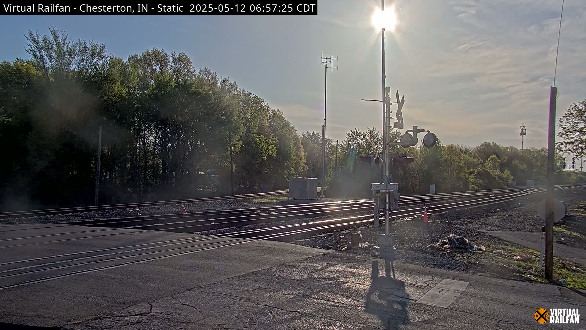 A sunny day at a railroad crossing shows several train tracks intersecting a road, with trees and a signal system visible.