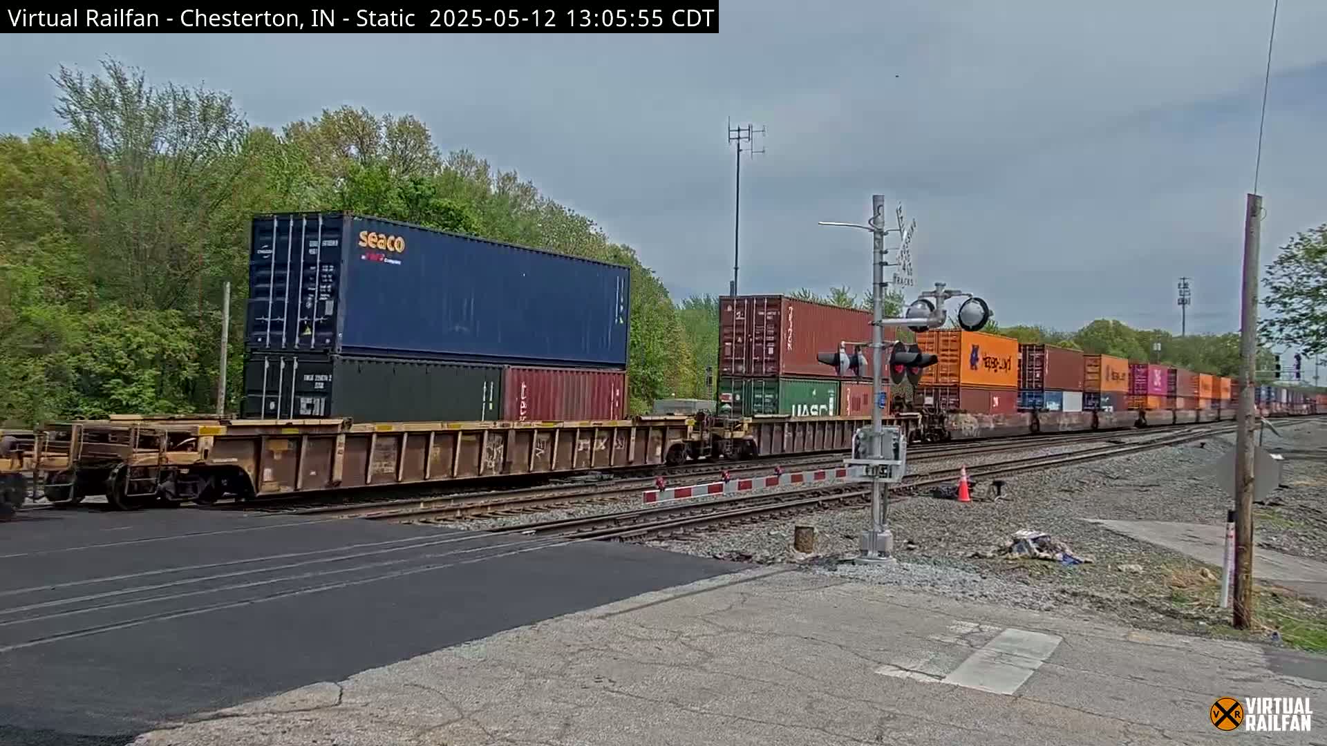 A long freight train carrying multiple shipping containers passes a road crossing under a partly cloudy sky.