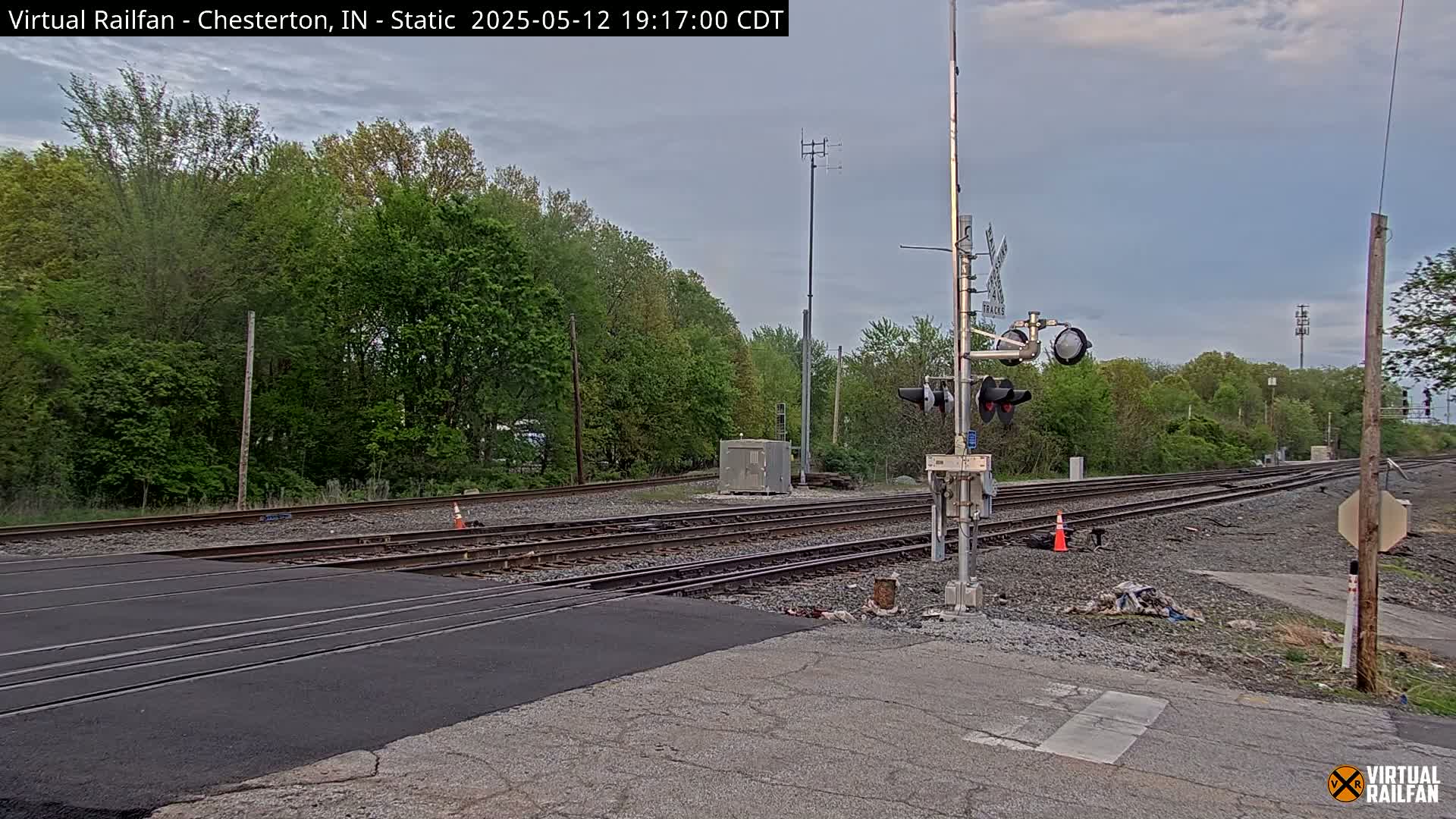 A railroad crossing with train tracks and a signal light is situated amidst lush green trees under a partly cloudy sky.