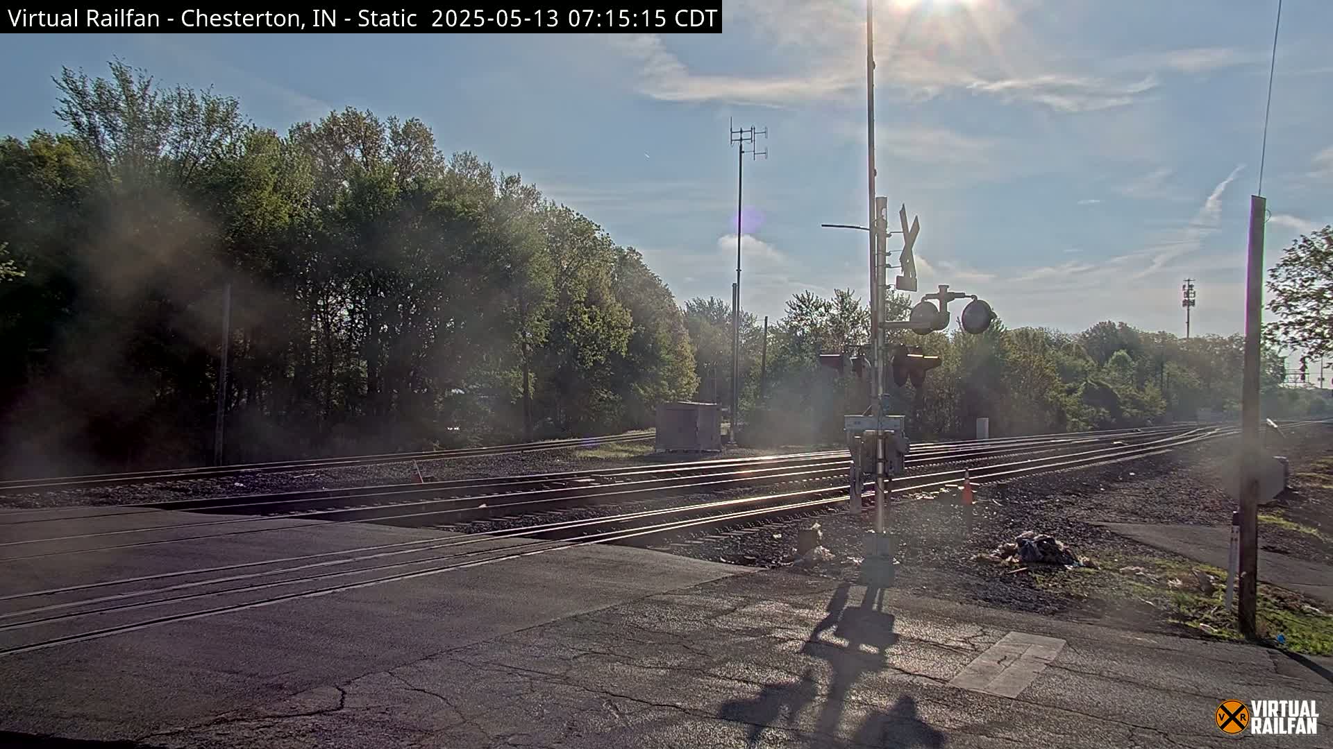 A sunny day reveals a railroad crossing with multiple tracks, signal lights, and a line of trees in the background, slightly obscured by a hazy atmosphere.