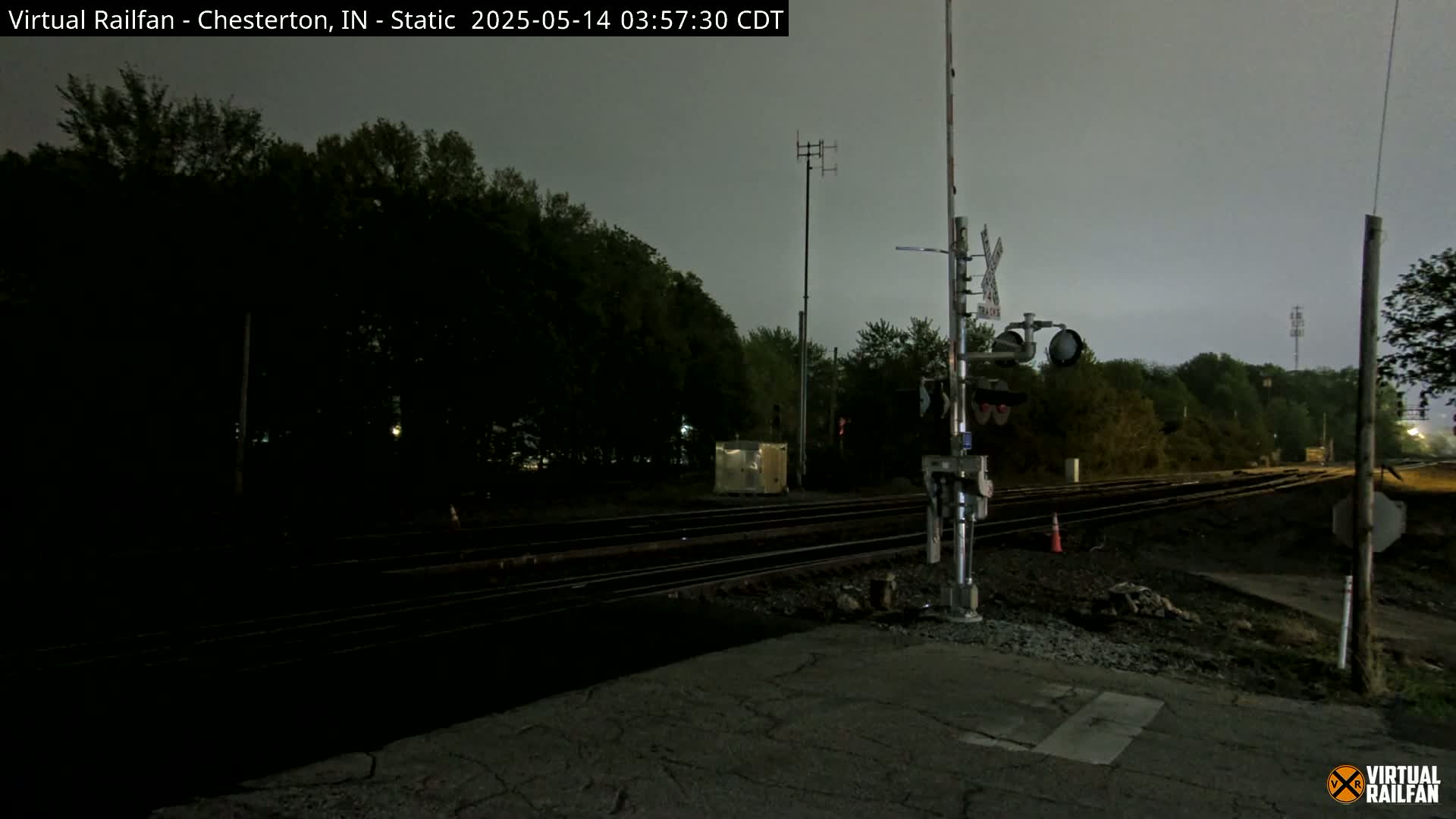 A nighttime view of a railroad crossing with train tracks, signals, and trees under an overcast sky.