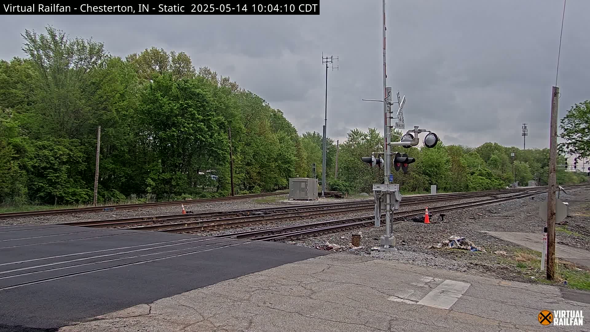 A railroad crossing with multiple tracks, railroad signals, and lush green trees under an overcast sky.