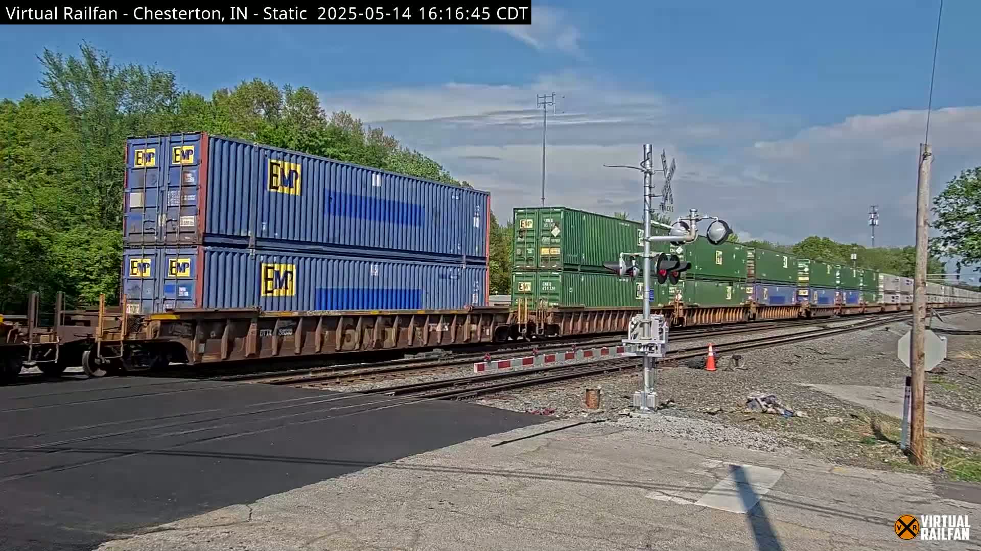 A freight train carrying stacked shipping containers passes a railroad crossing on a sunny day.