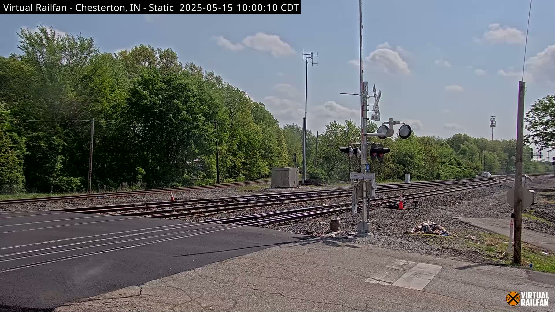 A railroad crossing with multiple tracks is shown, under a partly cloudy sky, surrounded by lush green trees.