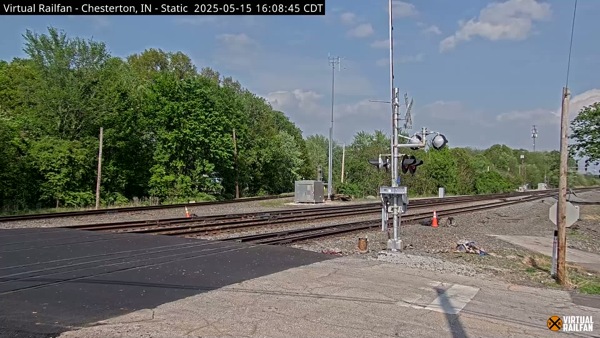 A railway crossing with a newly paved surface is shown under a partly cloudy sky, bordered by lush green trees.