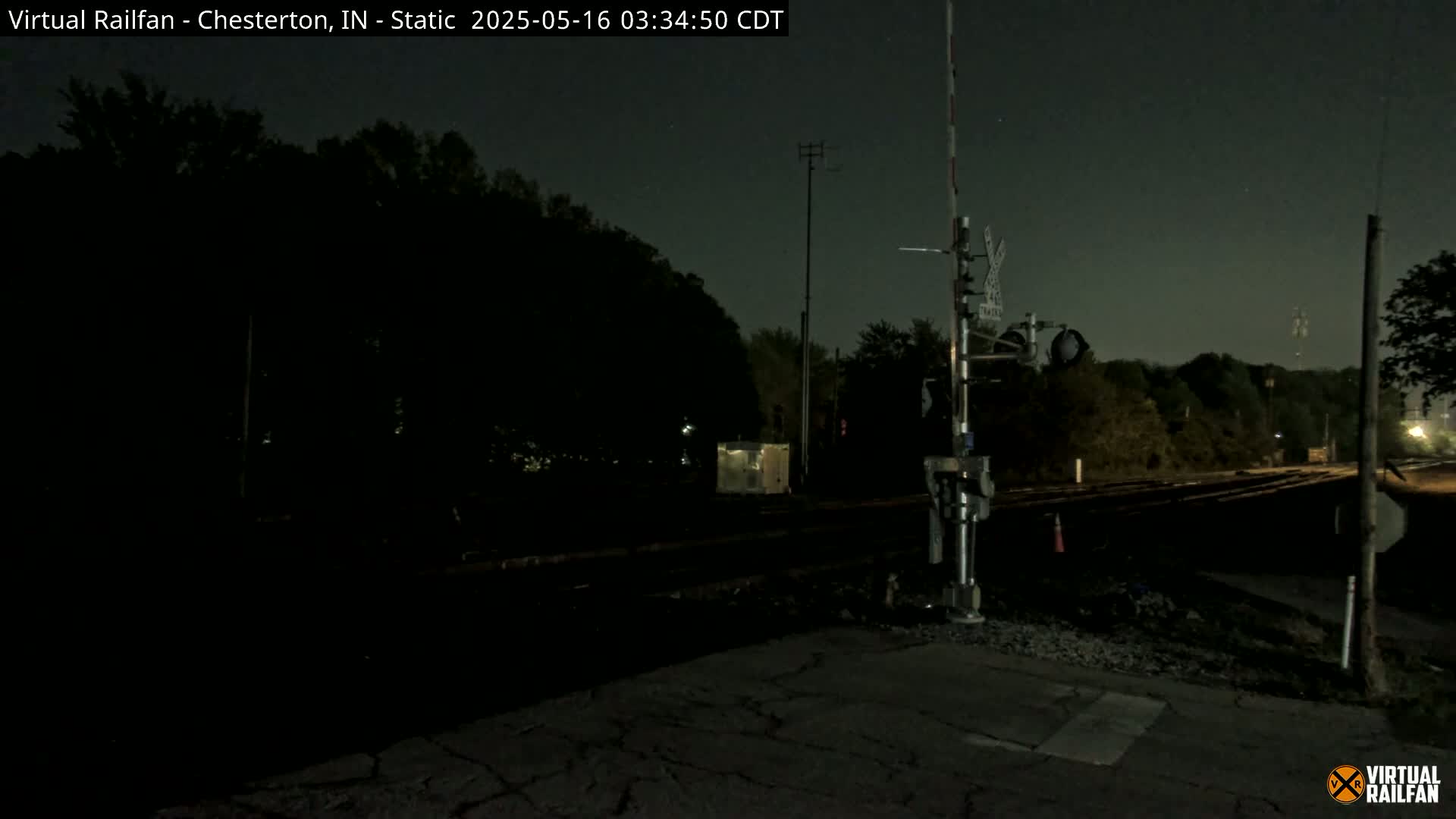 A nighttime view of a railroad crossing with a signal light and trees in the background under a dark sky.