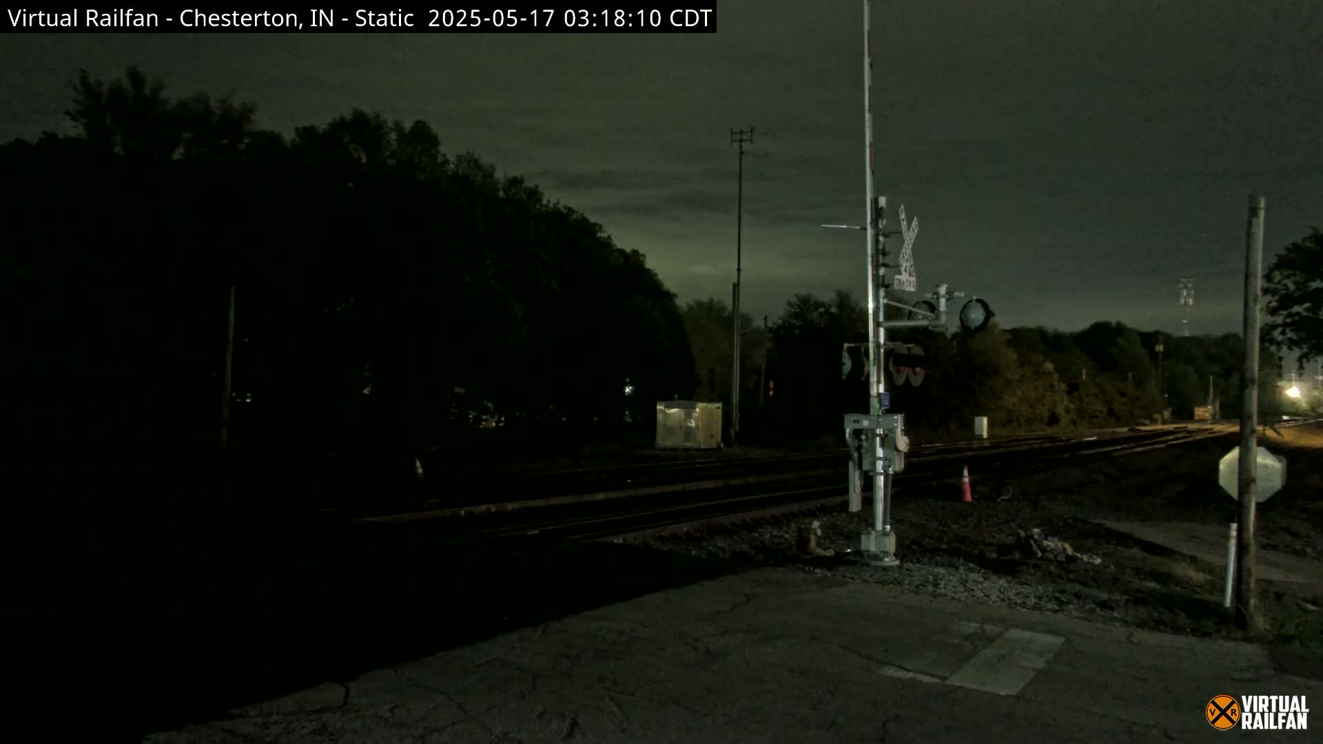 A nighttime view of a railroad crossing with train tracks extending into the dark, tree-lined distance under a cloudy sky.