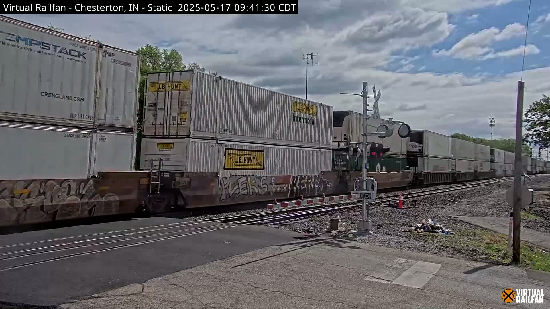 A long freight train carrying numerous white shipping containers passes a railway crossing under a partly cloudy sky.
