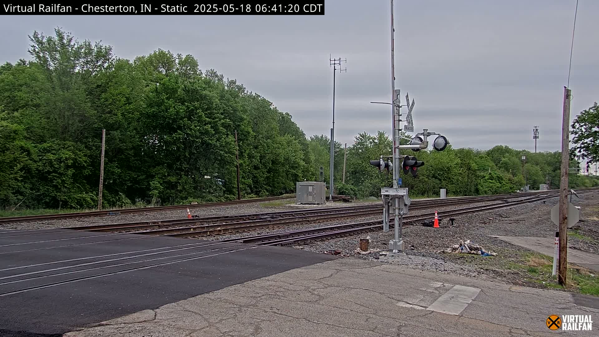 A railroad crossing with multiple tracks and a signal system is shown, surrounded by lush green trees under an overcast sky.