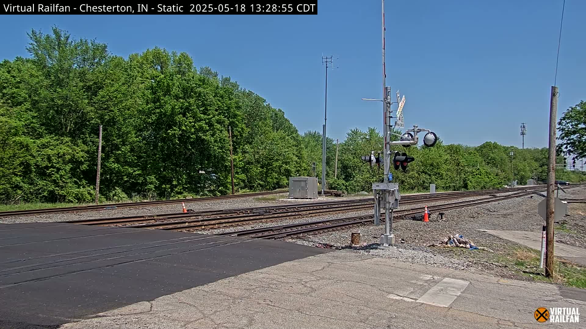 A railroad crossing with three sets of tracks is shown on a sunny day, surrounded by lush green trees.