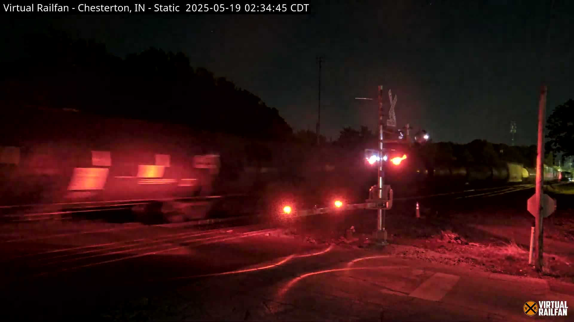 A train passes through a nighttime railroad crossing, illuminated by the red lights of the crossing gates.