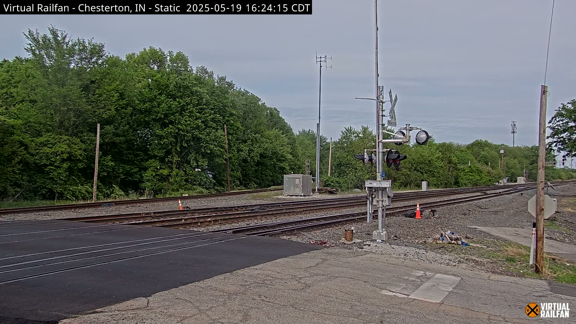 A railroad crossing with multiple tracks is seen under an overcast sky, surrounded by lush green trees.
