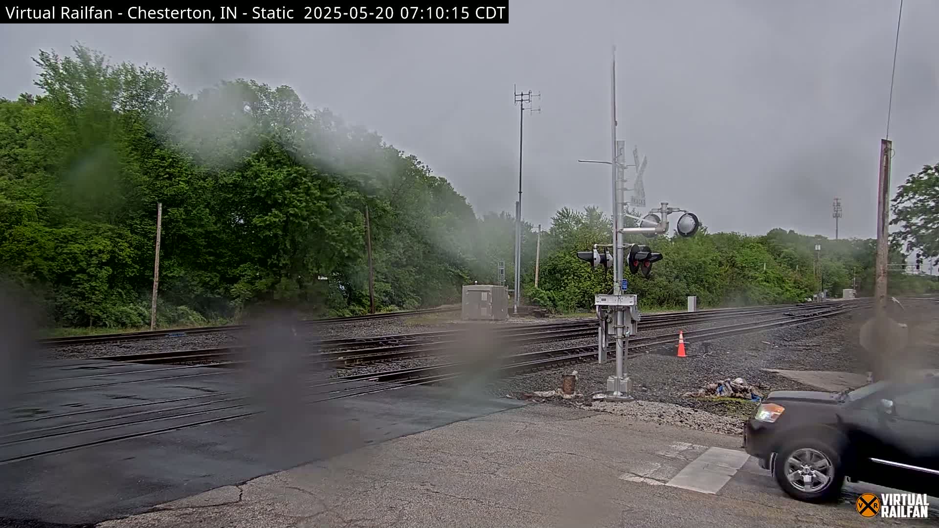 A rain-covered view of a railroad crossing with multiple tracks, surrounded by lush green trees, and a dark-colored pickup truck waiting at the crossing.
