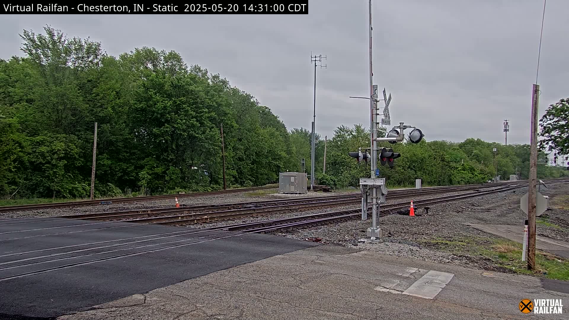 A railroad crossing with traffic signals is situated beneath an overcast sky, surrounded by lush green trees.