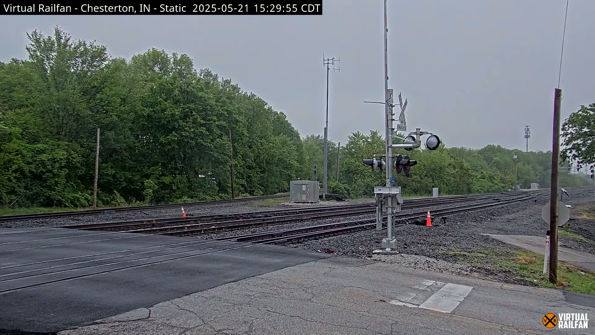 A railroad crossing with multiple train tracks and traffic signals is shown on an overcast day, surrounded by lush green trees.
