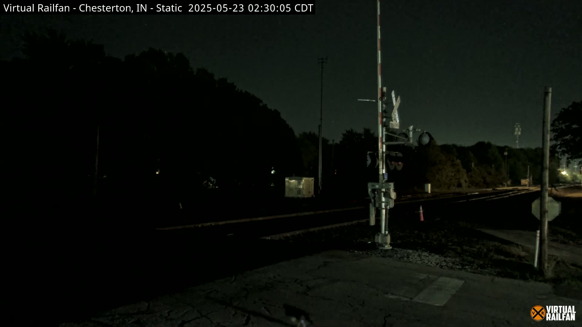 A nighttime view of a railroad crossing with a train signal, surrounded by dark trees under a clear sky.