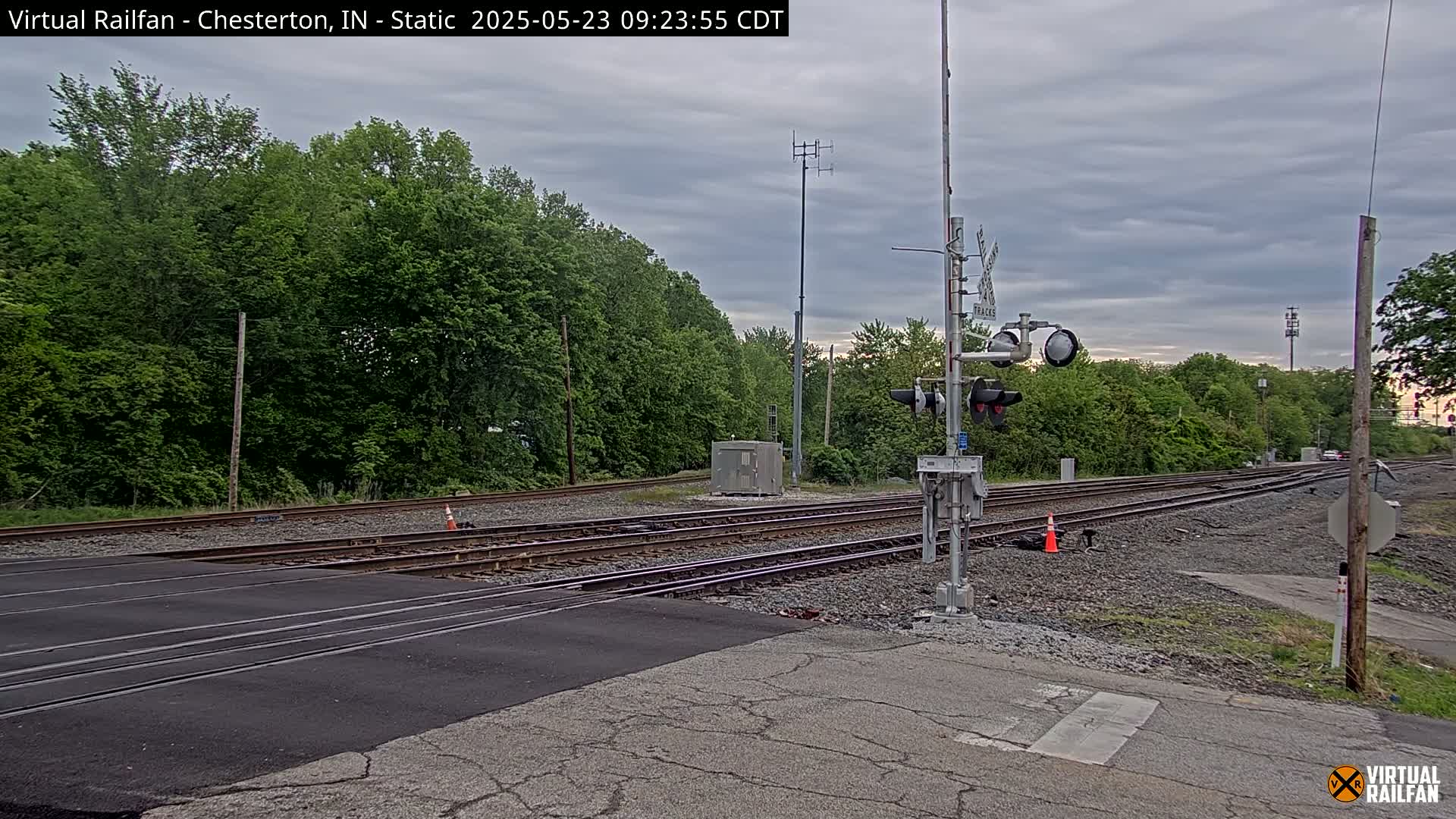 A railroad crossing with a traffic signal and multiple tracks is visible under an overcast sky, surrounded by lush green trees.