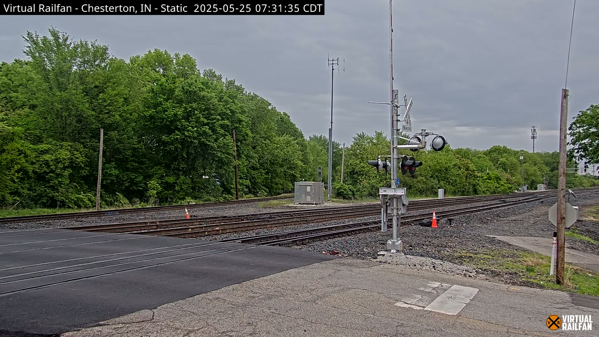 A railroad crossing with a traffic signal and several sets of tracks is visible under an overcast sky, surrounded by lush green trees.