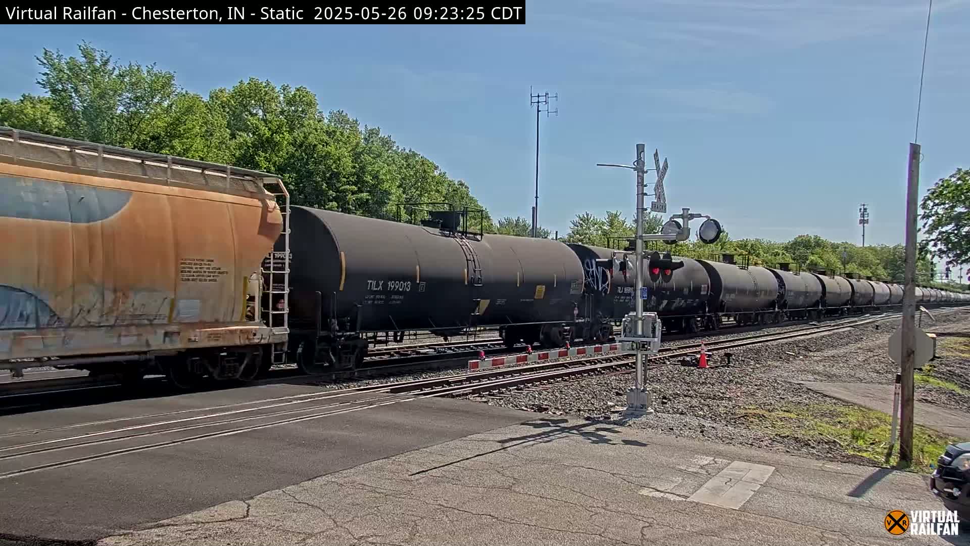 A long freight train of tanker cars passes a railroad crossing under a sunny sky.