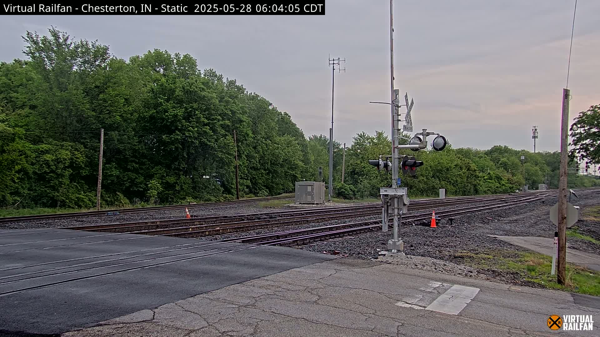 A railroad crossing with multiple tracks is seen under an overcast sky, surrounded by lush green trees.