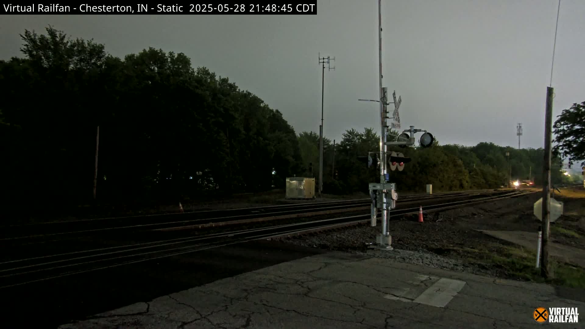 A nighttime view of a railroad crossing under an overcast sky shows train tracks leading into a wooded area, with a crossing signal and other equipment visible.