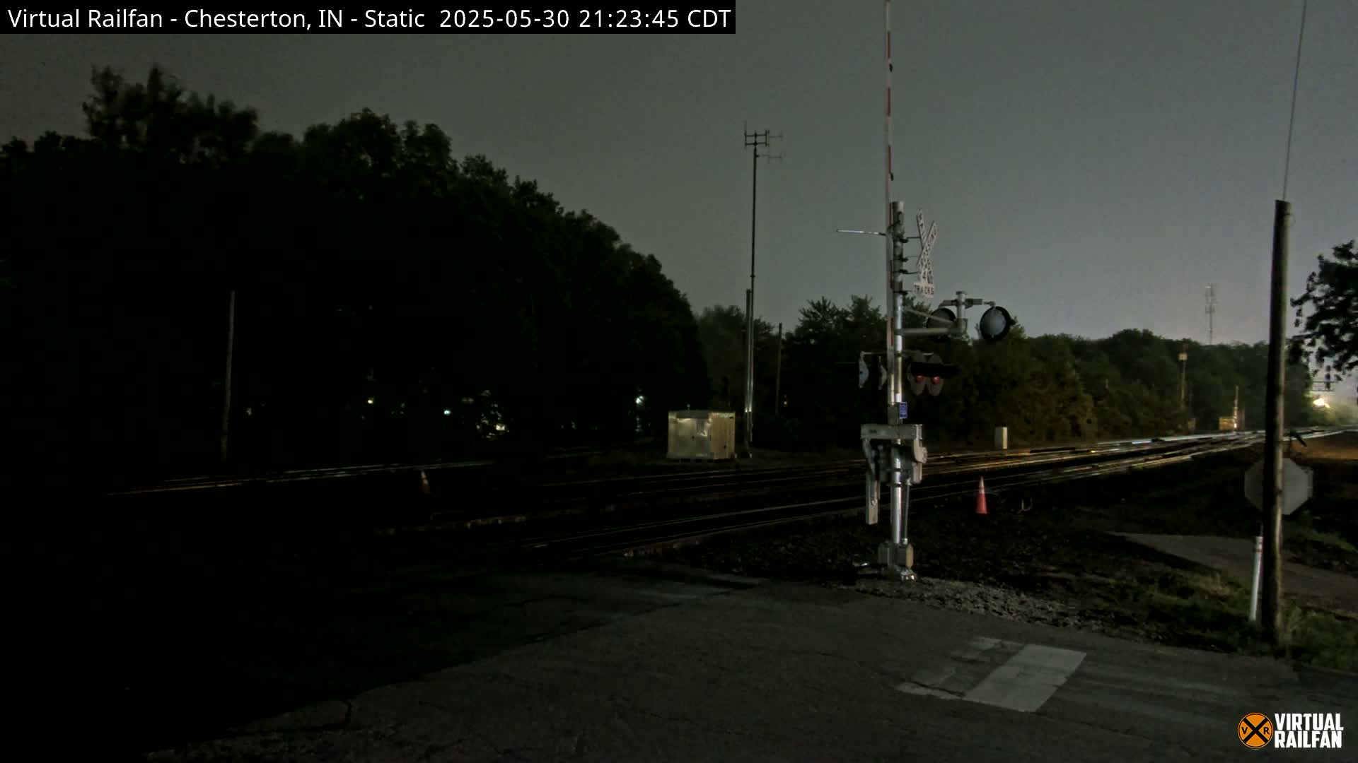 A nighttime view of a railroad crossing with train tracks, signals, and surrounding trees under a dark, overcast sky.