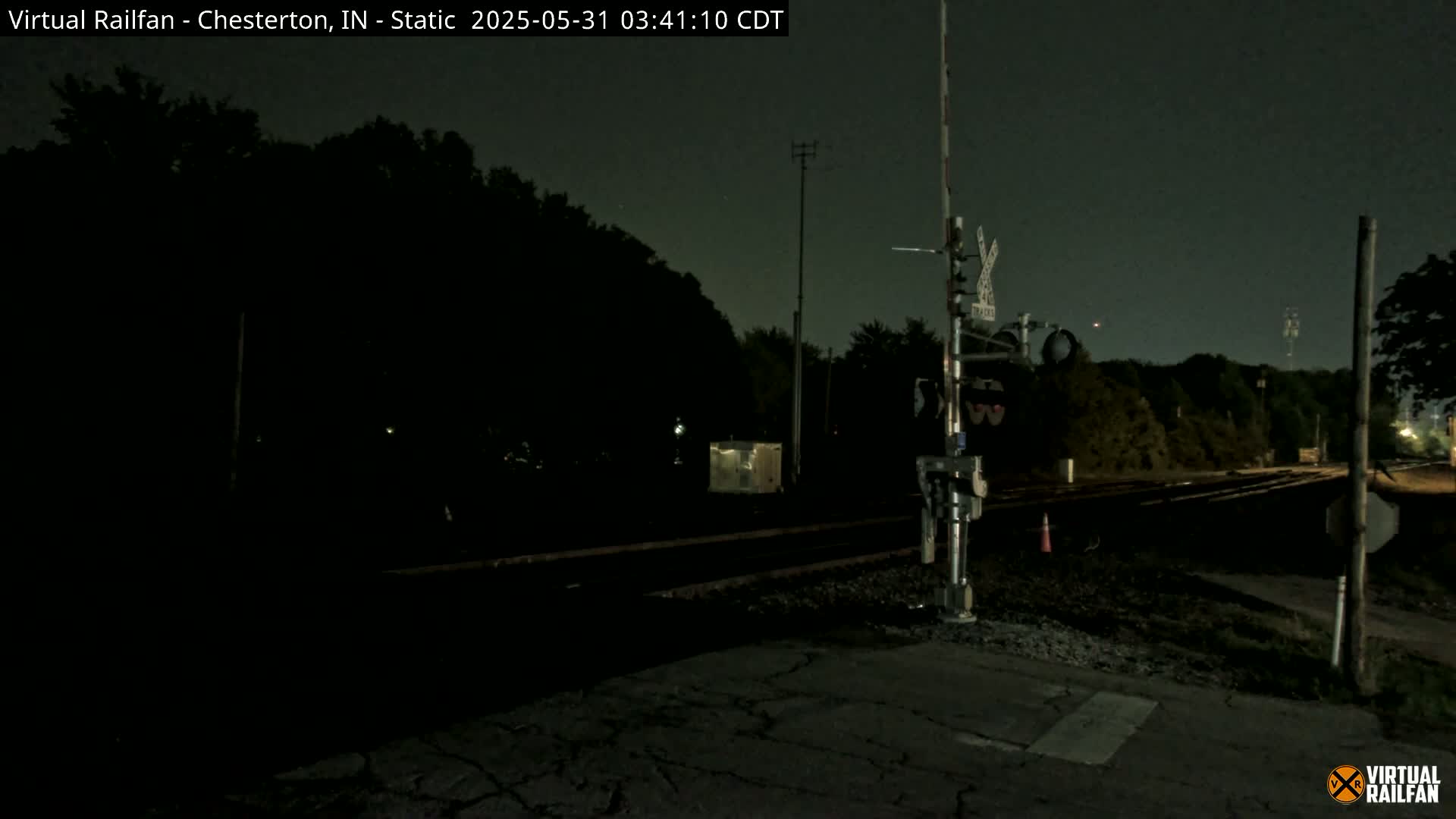 A nighttime view of a dark railroad crossing with a signal light, trees, and a roadway.