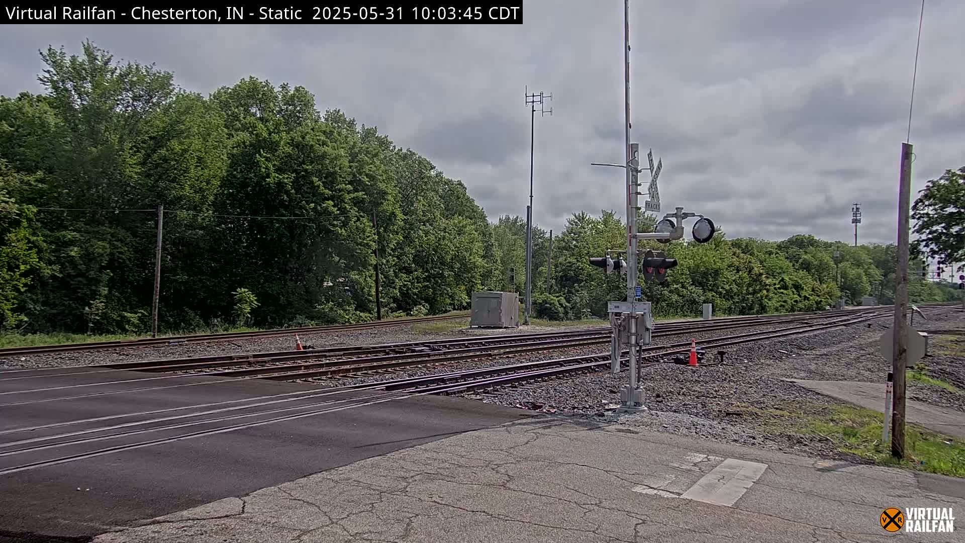 A railroad crossing with multiple tracks is seen under a partly cloudy sky, surrounded by lush green trees.