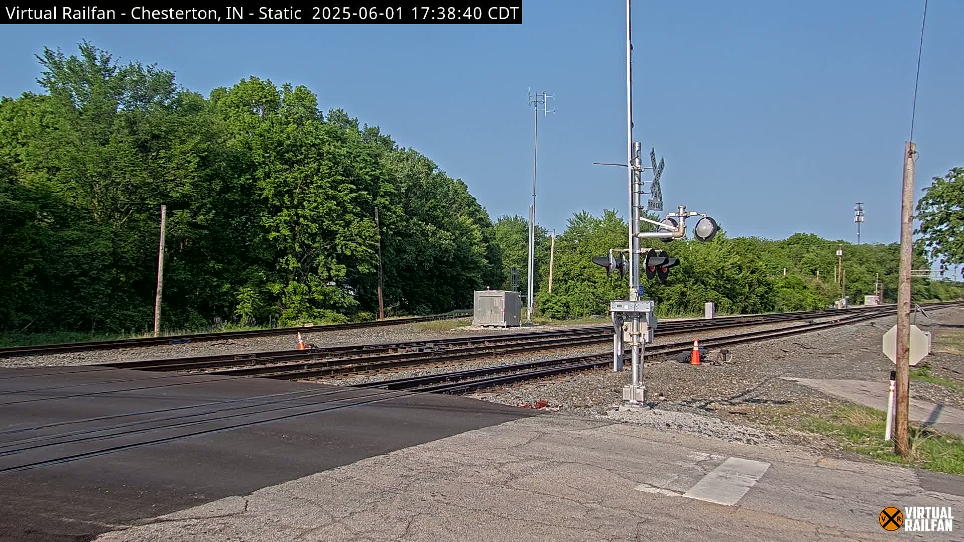 A railroad crossing with a signal light and multiple tracks is surrounded by lush green trees under a clear blue sky.