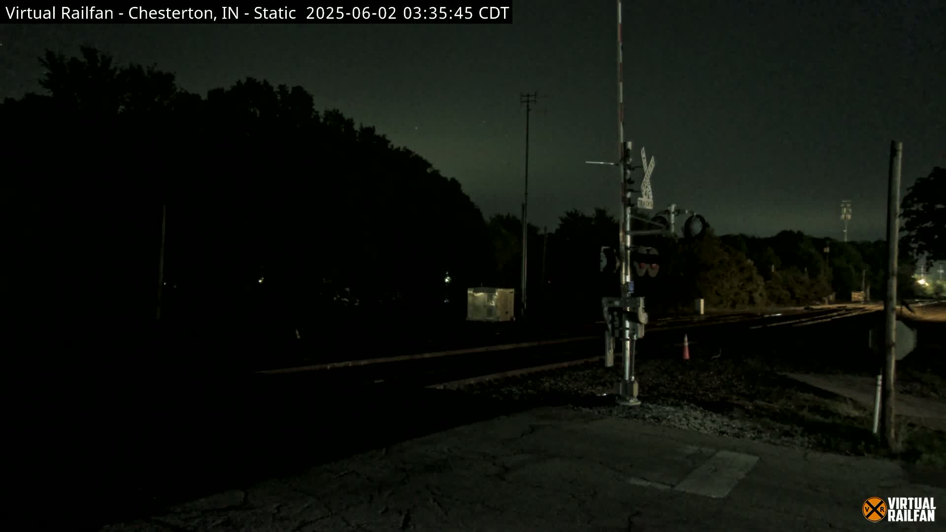 A nighttime view of a railroad crossing with a signal, surrounded by dark trees under a clear, dark sky.