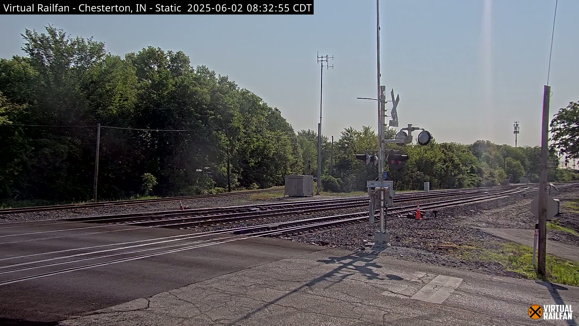 A sunny day at a railroad crossing shows multiple train tracks intersecting a road, with lush green trees in the background and railway signals in place.