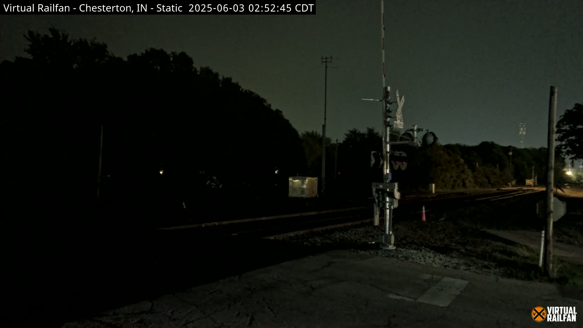 A nighttime view of a dark railroad crossing with a signal light and trees in the background under a cloudy sky.
