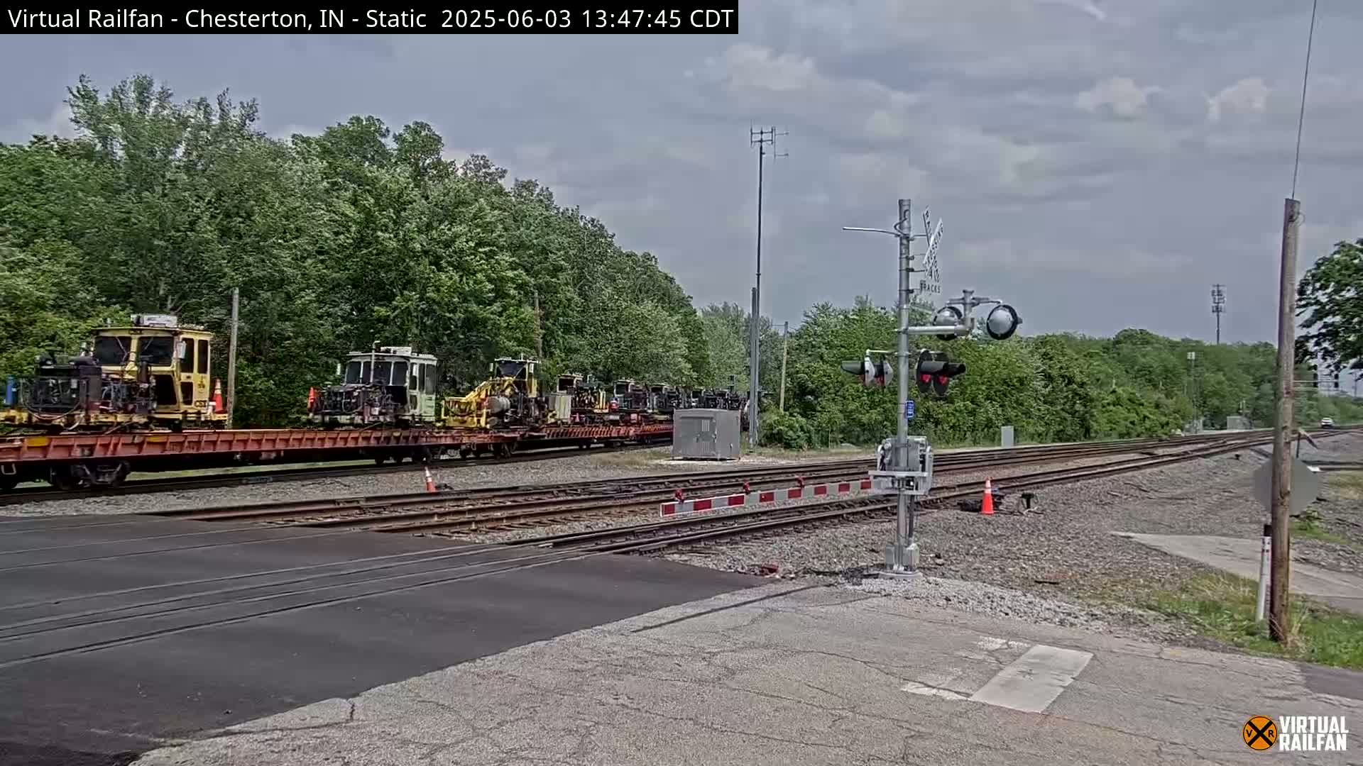 A flatbed train car carrying several pieces of yellow and green construction equipment sits on a railway crossing under a partly cloudy sky.