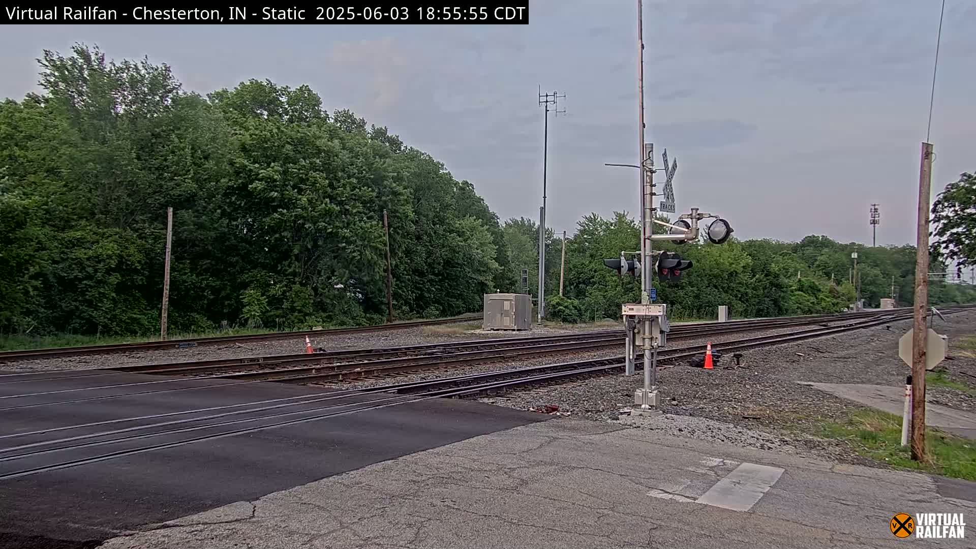 A railroad crossing with a road intersecting two sets of train tracks under an overcast sky, surrounded by lush green trees.