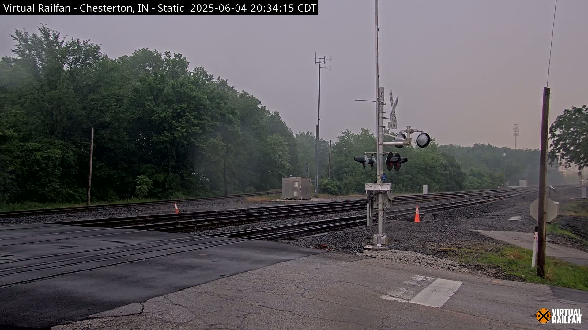 A railroad crossing with multiple tracks is seen under an overcast sky, surrounded by lush green trees.