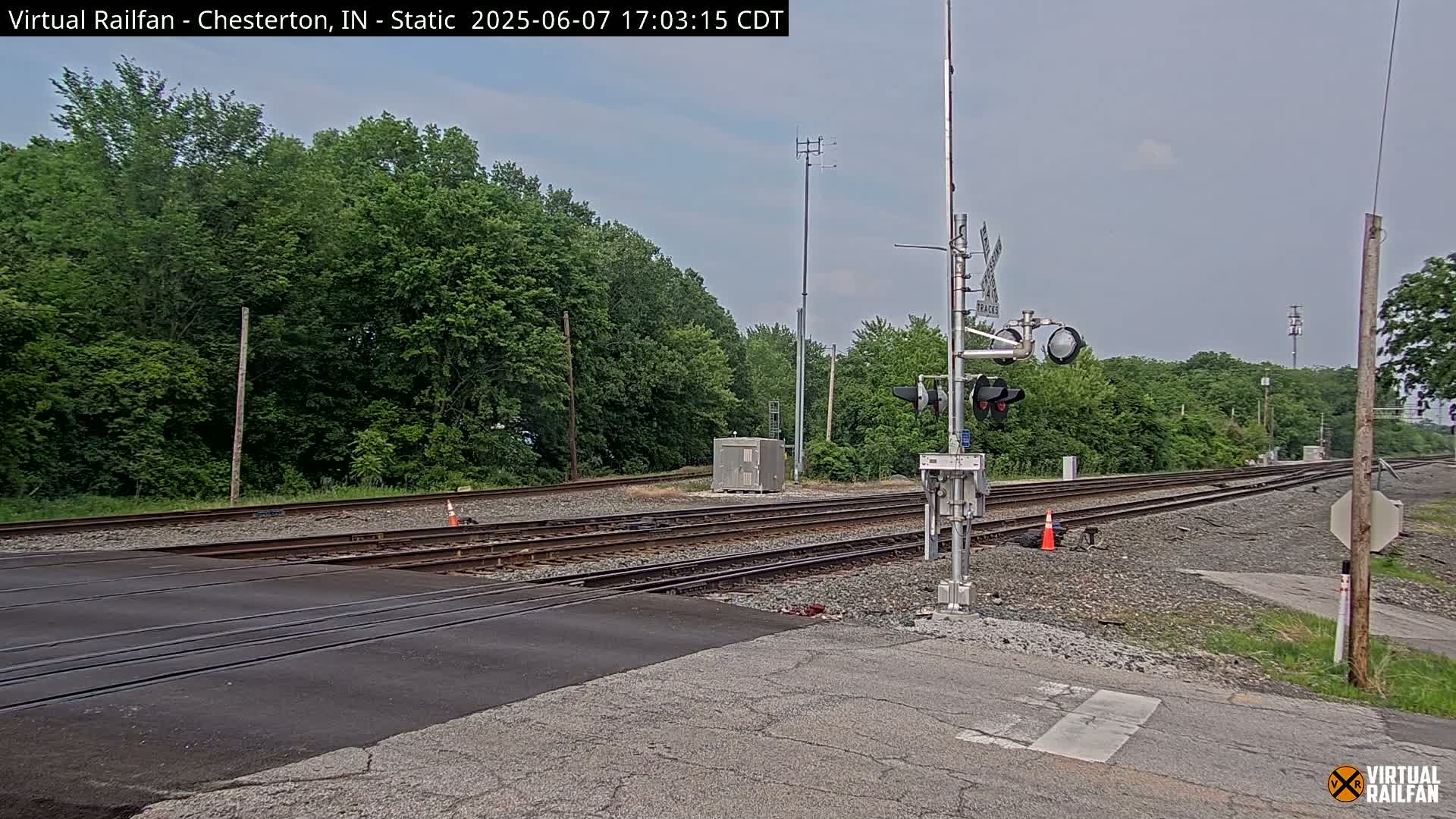 A railroad crossing with multiple tracks is situated near a lush green wooded area under an overcast sky.