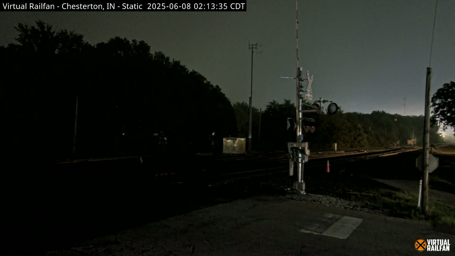 A nighttime view of a railroad crossing with dark trees in the background under an overcast sky.