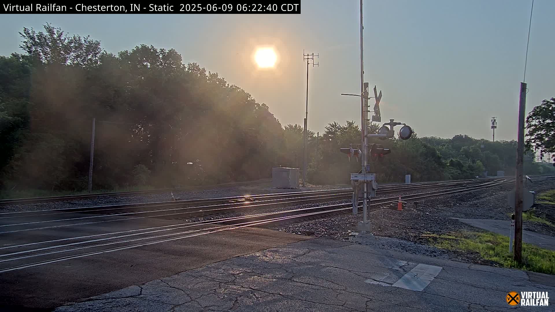 A sunlit railroad crossing with multiple tracks is surrounded by trees and hazy morning air.