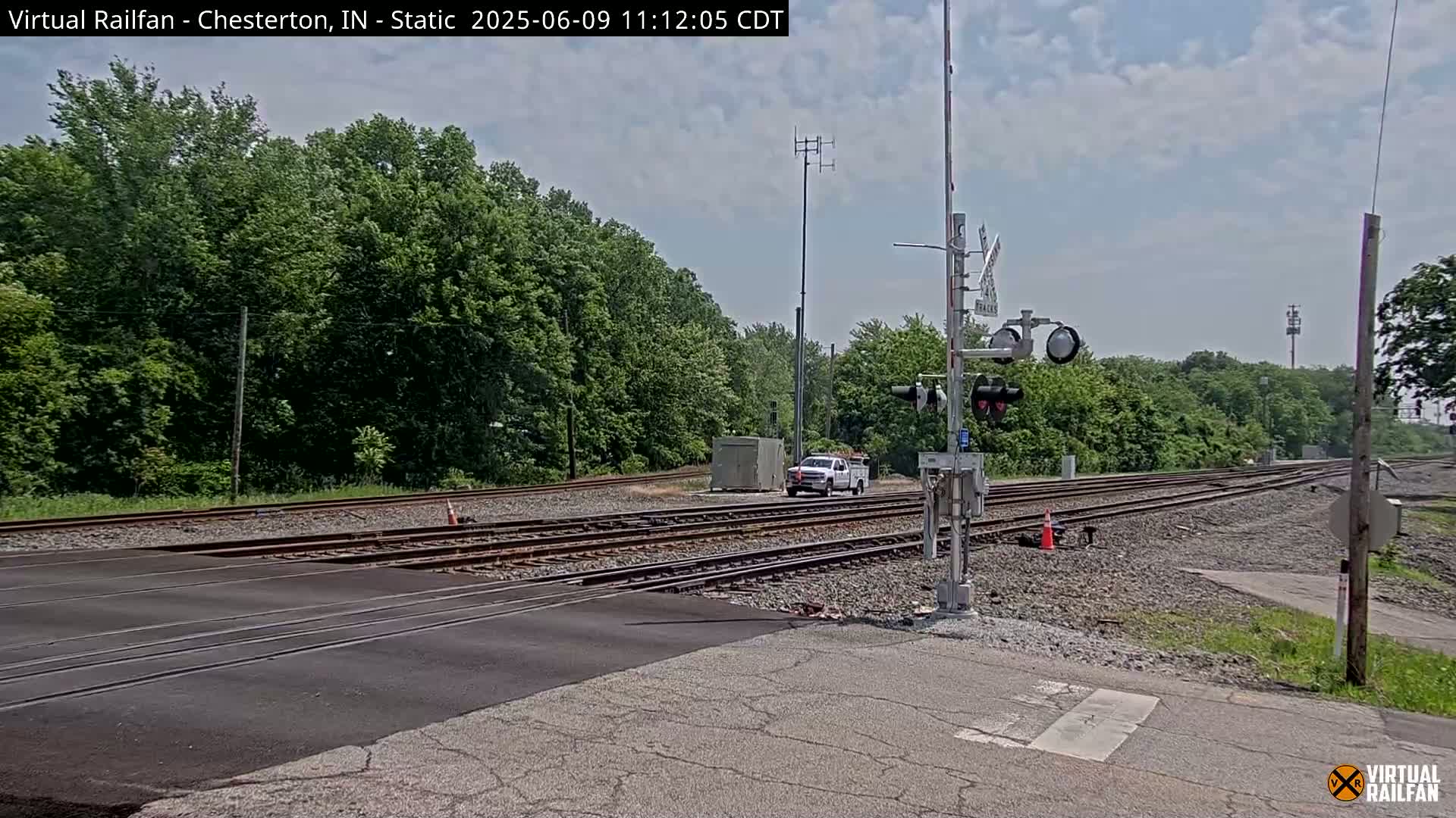 A white pickup truck is parked near a railroad crossing on a partly cloudy day, with lush green trees in the background.