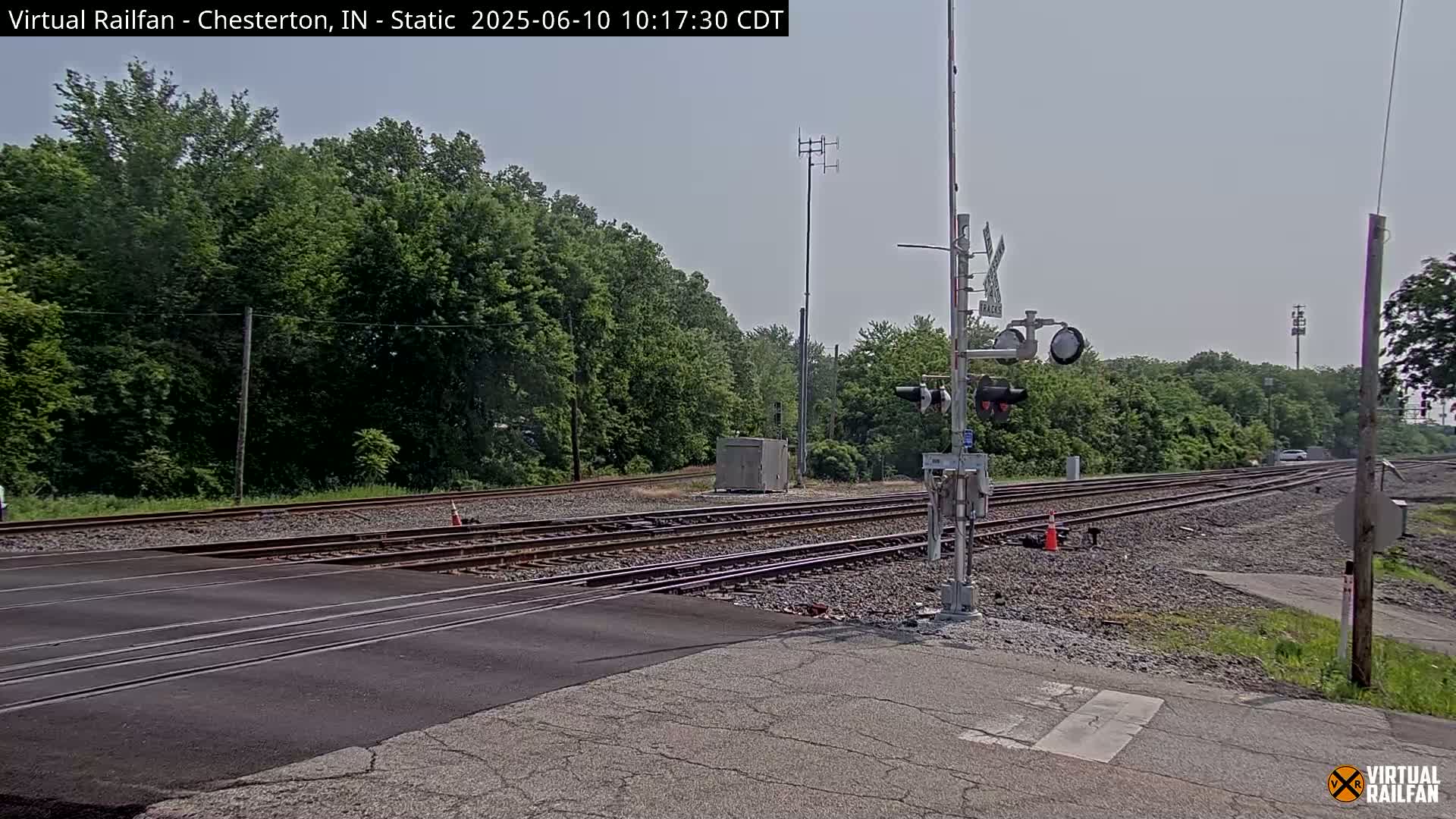 A road crosses over multiple sets of railroad tracks on a sunny day, with lush green trees lining the background.